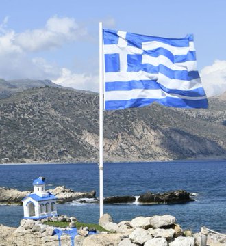 The blue and white Greek flag flies on a flagpole at a coastal location, with a small blue and white chapel and mountains visible in the background.