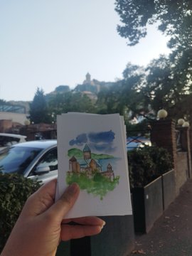 A hand holds a small watercolor painting of houses and trees in front of a real-life landscape with a domed building in the distance.