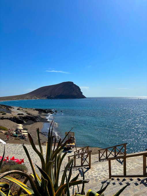 A scenic coastal view features a prominent rocky headland jutting into turquoise waters under a clear blue sky, with a sandy beach, wooden railings, and an agave plant in the foreground. The image has no connection to the accompanying post about "GM LEGENDS" and family, as it simply depicts a peaceful seaside landscape with no visible text or people in focus.