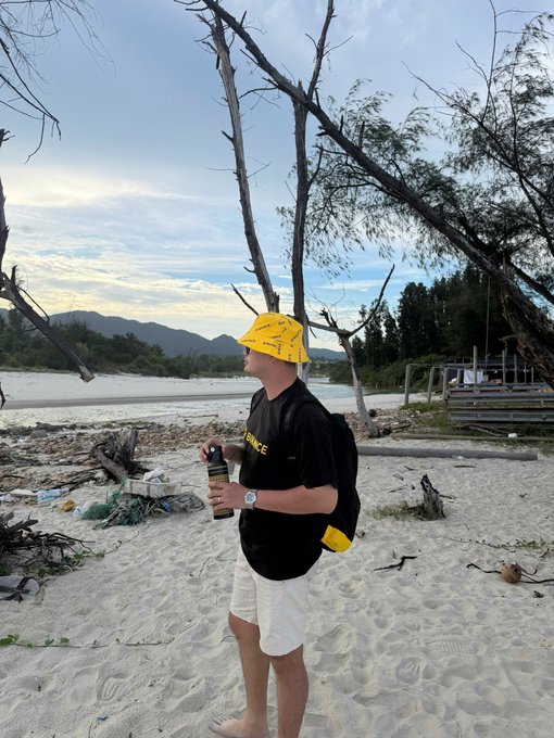 A man wearing a yellow hat, black 'BINANCE' t-shirt, and white shorts stands on a sandy beach holding a bottle, with mountains and bare trees in the background.