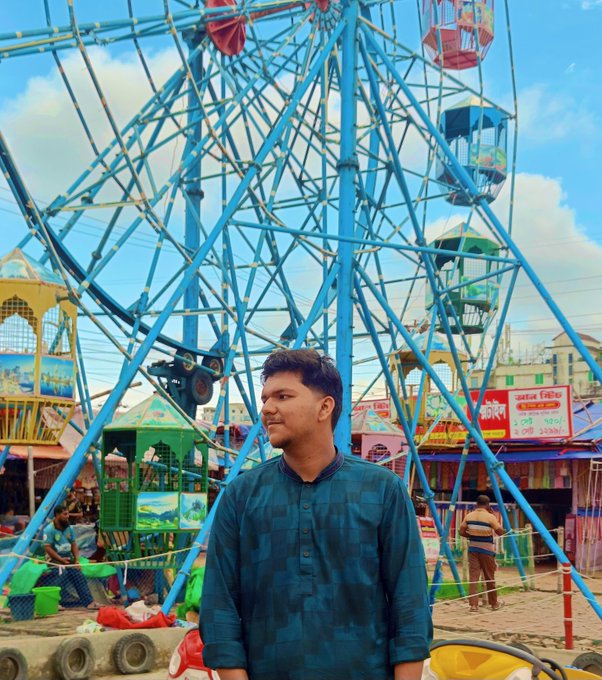 A young man in a dark teal traditional kurta standing in front of a blue Ferris wheel at what appears to be a fair or festival.