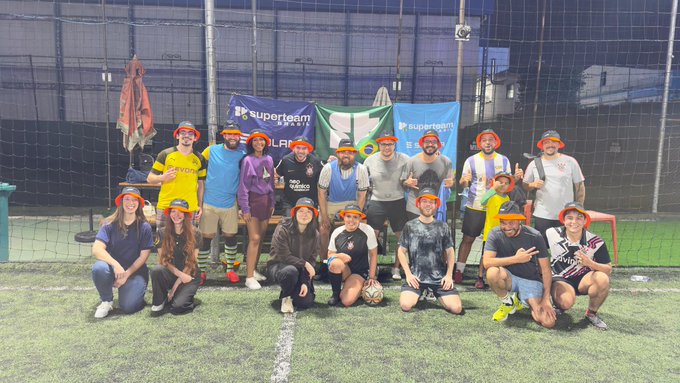 A group of around 15 people wearing matching red and black headbands pose for a team photo on an indoor soccer field, with a soccer ball at center and banners reading "Superteam BR" and "LFG" behind them—capturing the crossover moment where the Cashi and SuperteamBR builders traded their laptops for the pitch in Brazil.