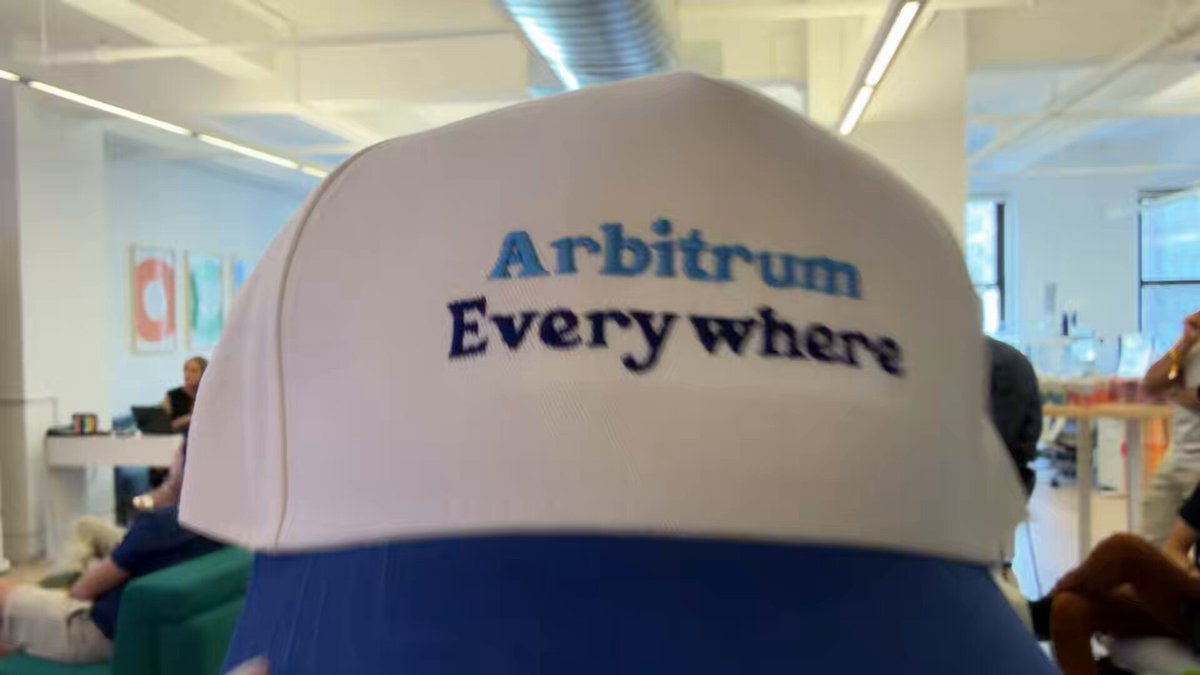 The image shows a close-up of a white baseball cap with blue trim, which has the words "Arbitrum" written on it in black letters. The cap appears to be worn backwards or tilted slightly, giving it an unusual appearance. In the background, there are people present, but they are blurred and not the main focus of the image.