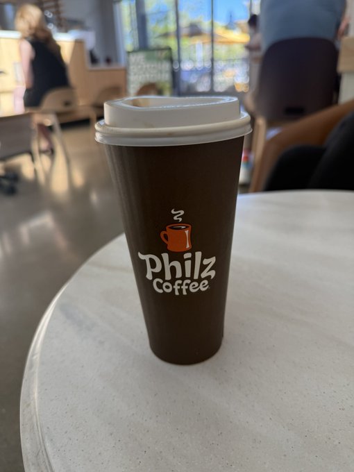 A tall brown "Philz Coffee" cup sits on a round marble table inside a coffee shop, with the brand's logo and steaming coffee mug icon clearly visible. The image pairs with the post's quip about crypto bears facing liquidation before the bulls have even finished their morning coffee.