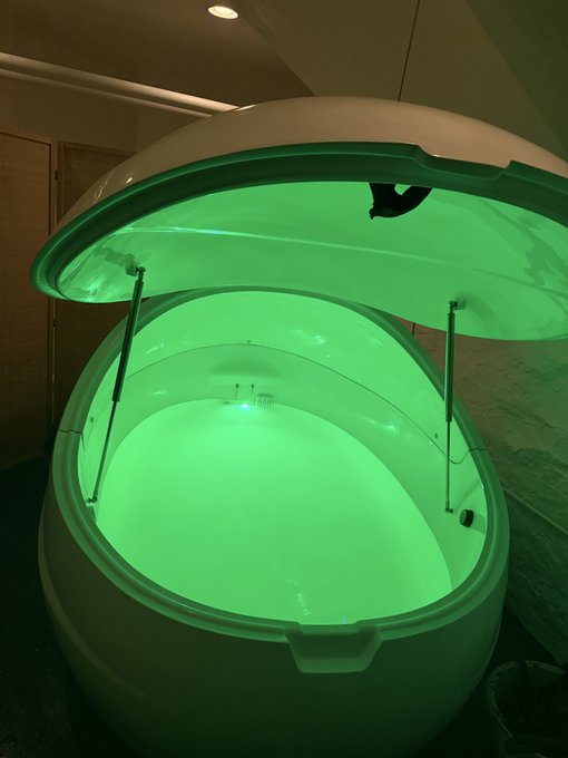 An open pod or capsule device illuminated with green light, possibly a sensory deprivation or flotation tank, sits in a dimly lit room.