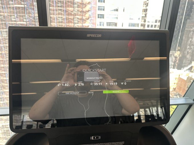 A Precor gym treadmill display screen showing 'QUICKSTART' workout paused, with stats 1.82km, 276 calories, 35:11 time, 19:19 pace, 0 BPM heart rate, and 'END WORKOUT' and 'RESUME' buttons, reflecting a person taking a photo.