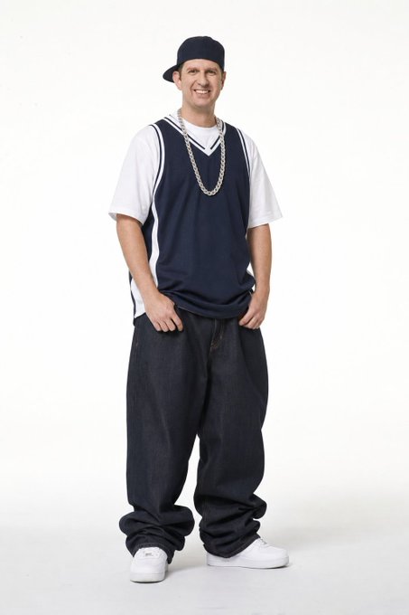 A man poses in a backwards baseball cap, oversized chain, baggy basketball jersey over a white tee, and very wide-legged dark jeans, channeling an early 2000s hip-hop aesthetic reminiscent of Eminem's iconic style.