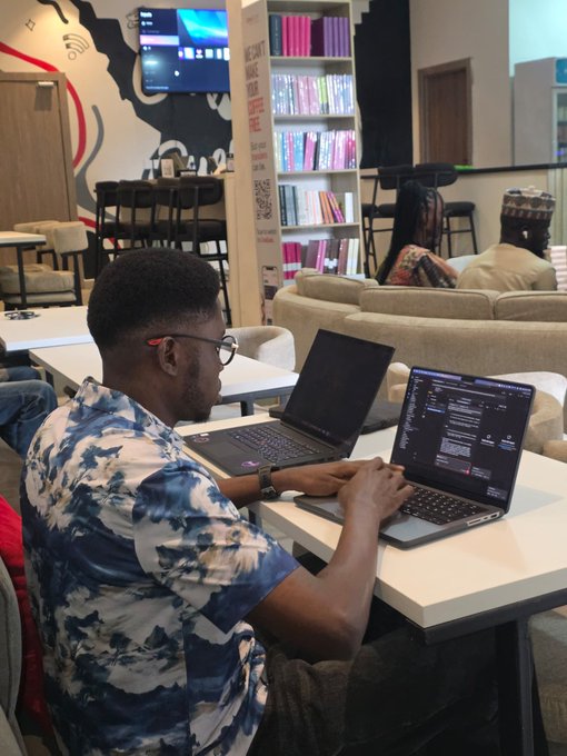 A casual photo of a man working on two laptops at a table in what appears to be a modern office or co-working space, with other people and a bookshelf visible in the background.