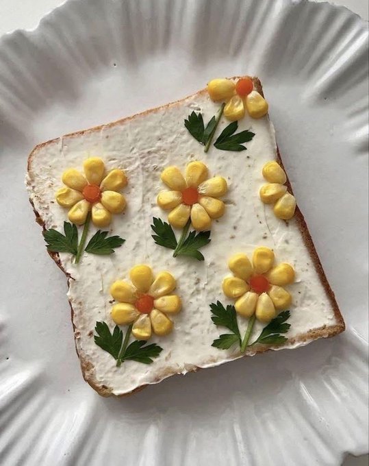 A slice of toast on a white ridged plate, spread with cream cheese or mayo and decorated with corn kernels arranged as yellow daisy flowers with orange centers, accented by parsley leaves.