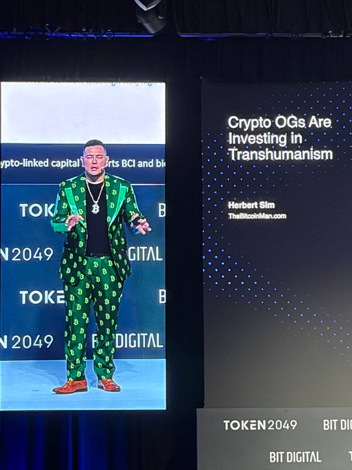 A man wearing a green patterned suit and orange shoes speaks on stage at TOKEN2049 BIT DIGITAL, standing before a backdrop with the slide title 'Crypto OGs Are Investing in Transhumanism' by Herbert Sim of TheBitcoinMan.com.