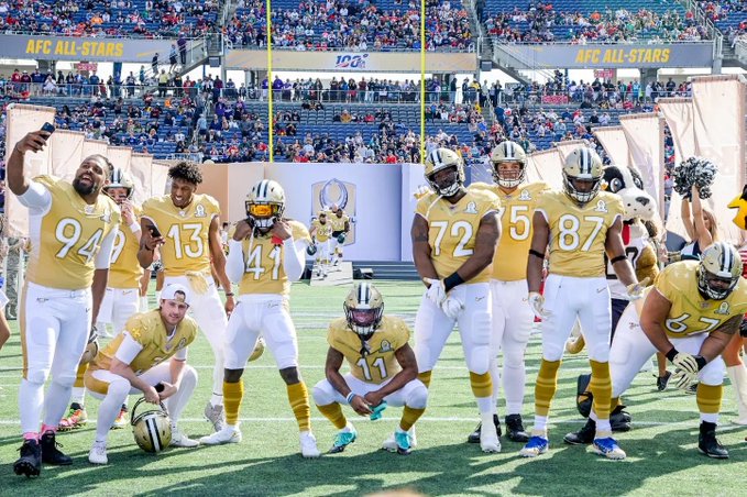 Football players in gold and white AFC All-Stars uniforms pose for a group photo on the field, with stadium seating and "AFC ALL-STARS" banners visible in the background.