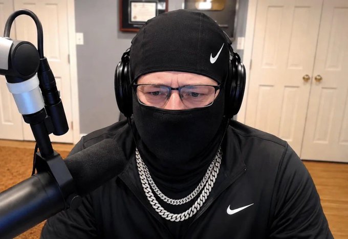 A person sits in front of a microphone wearing a black Nike skull cap, black balaclava covering the lower face, headphones, glasses, and a thick silver chain, giving off a trapper aesthetic. The masked, jewelry-heavy look directly plays into the post joking about when @anonchain started "trappin'."