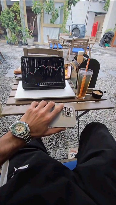 A first-person view of someone wearing a silver watch and Puma sweatpants, using a smartphone with an iPad displaying trading charts and a tall glass of iced tea on a wooden table at an outdoor cafe.