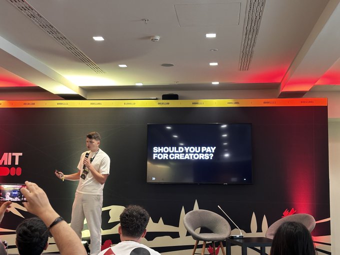 A man speaks on stage at a conference, with a screen behind him displaying the question 'SHOULD YOU PAY FOR CREATORS?'