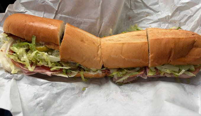 A large, footlong submarine sandwich cut into sections, loaded with deli meat, shredded lettuce, and pickles on a soft hoagie roll, rests on white wrapper paper with a partial "Jersey Mike's" logo visible. The image perfectly illustrates the post's praise for a giant sub from Jersey Mike's, showcasing the sandwich's generous size and fresh toppings.