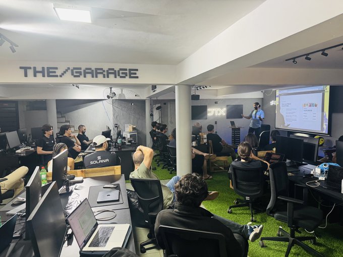 An interior view of a tech/crypto workspace called 'THE GARAGE' where a group of people are gathered around desks with monitors, watching a presentation displayed on a large screen.