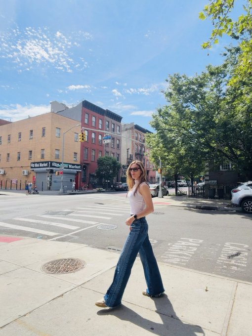 The image shows a woman walking down a city street, wearing jeans and sunglasses. She appears to be enjoying her time outdoors. The surrounding area includes tall buildings, indicating that she is in an urban setting. There are also other people present on the sidewalk, suggesting that this location is a popular spot for pedestrians.