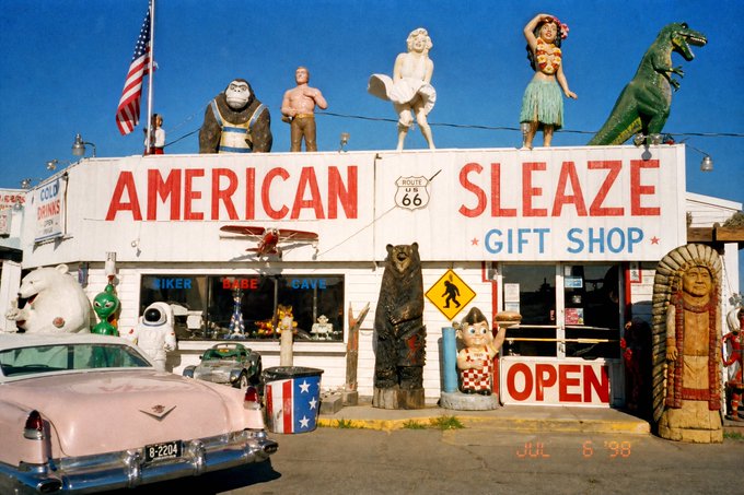 The image shows the "American Sleaze Gift Shop" along Route 66, decorated with eclectic roadside attractions including a dinosaur, Marilyn Monroe, hula girl, gorilla, and other kitschy statues, with a vintage pink Cadillac parked out front and a timestamp reading "JUL 6 '98." 

The image does not relate to the post's content about an NFT drop called "Holy" on Verse and a website exaltedlove.com—the photo depicts a quirky Route 66 tourist shop rather than any digital artwork, collection release, or promotion tied to the post.
