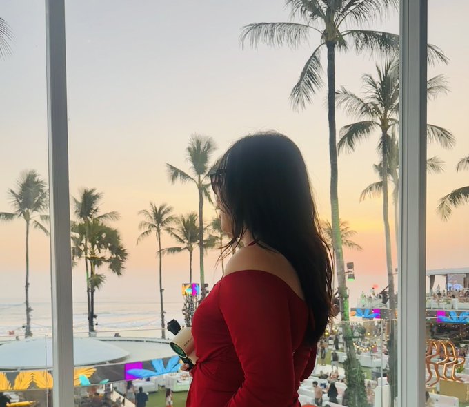 A woman in a red off-shoulder dress and sunglasses looks out a window at a sunset over a beach with palm trees and a crowded boardwalk.