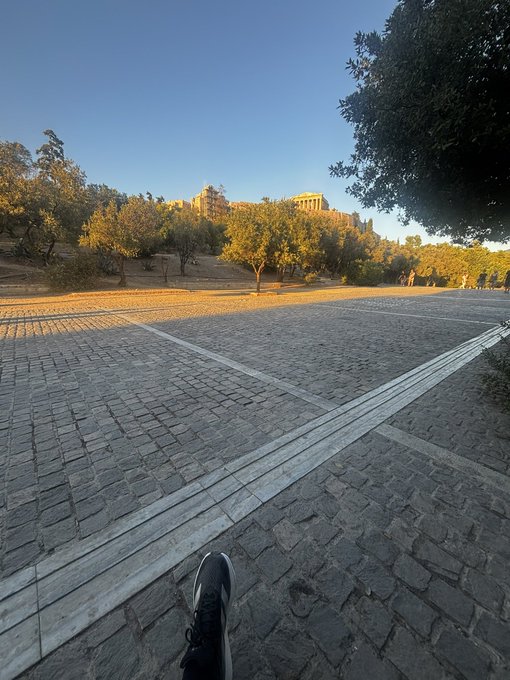 A first-person view of cobblestone pavement with a single sneaker visible, leading toward the Parthenon atop a hill in Athens under a clear blue sky.