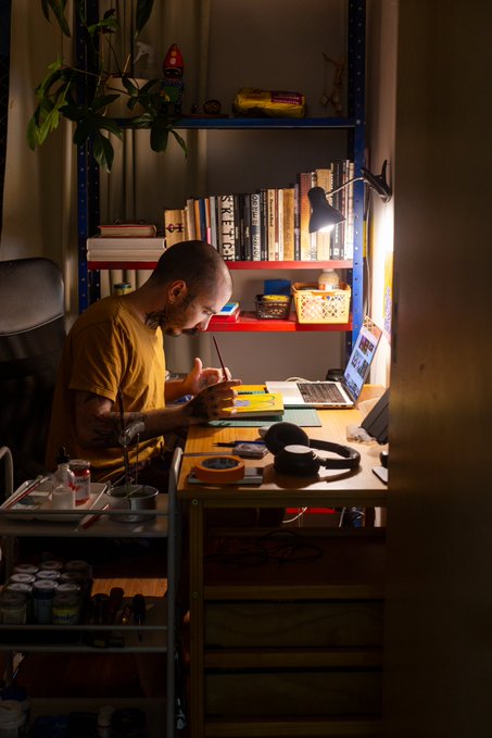 A tattooed man in a yellow shirt works at a desk with a laptop and drawing tablet, illuminated by a desk lamp in a room with blue shelving and plants.