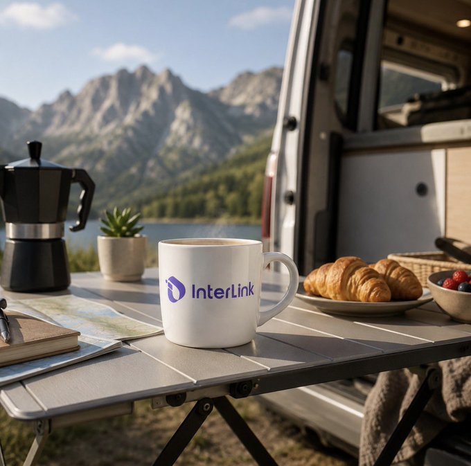 A camping scene with an "InterLink" branded mug on a table alongside croissants, coffee maker, and notebook, with mountains and a van in the background.