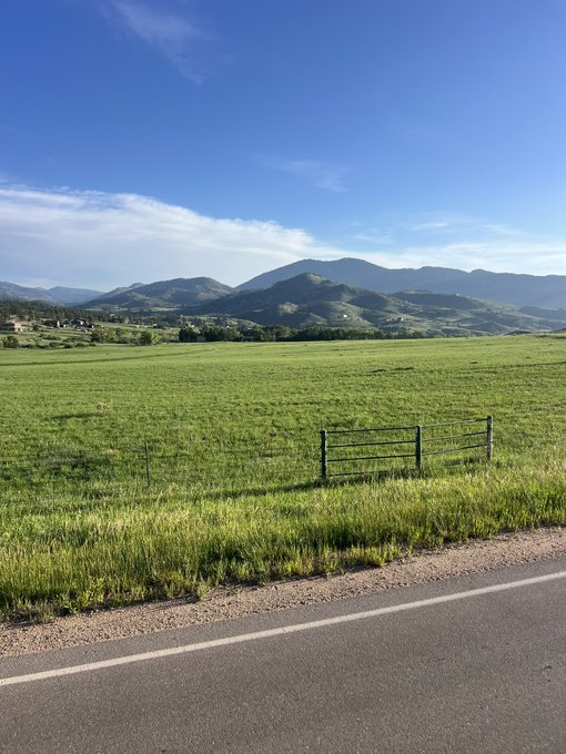 The image shows a large, open field with a green fence running along its edge. In the background, there are mountains visible against a clear blue sky. The photo appears to be taken from the side of a road or highway, as indicated by the presence of a white line on the ground and the perspective of the photograph.