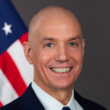 A smiling, bald man in a dark suit stands before an American flag in an official portrait. Based on the accompanying news, the image is being used to depict Sam Waldon, the SEC's Principal Deputy Director of Enforcement who is departing the agency after 14 years of service.