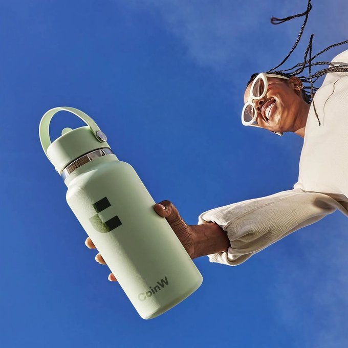 A smiling woman with white sunglasses and long braids holds up a sage green water bottle branded "CoinW" against a vibrant blue sky. The image visually reinforces the post's message by pairing hydration with the CoinW brand, suggesting that staying hydrated complements a successful, profitable lifestyle.