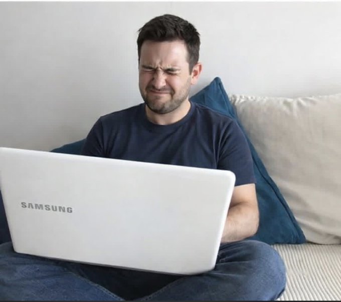 The image shows a man sitting on a couch with his laptop open, looking at something on the screen. He appears to be checking his long-term crypto investments or researching about them. The setting suggests that he might be working from home or staying updated on financial news and trends related to cryptocurrency.