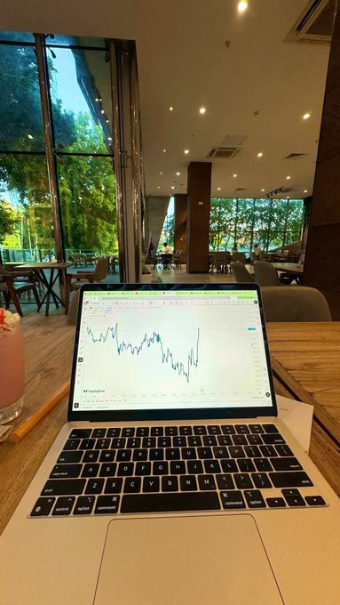 The image shows an open laptop computer with a graph displayed on its screen. The laptop is placed on top of a wooden table, which also has a drink next to it. In the background, there are several chairs and dining tables arranged around a large window that offers a view of trees outside. A person can be seen sitting at one of these tables in the distance. The scene suggests an office or workspace setting where people might be working on projects related to finance or data analysis.