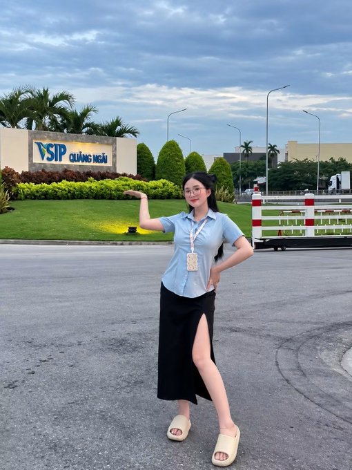 A young woman wearing glasses, a light blue shirt, and a black slit skirt poses with her arm extended in front of a 'VSIP Quảng Ngãi' sign and landscaped entrance.