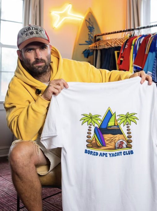 A man in a yellow hoodie and a 'Salty Crew' cap holds up a white t-shirt featuring a 'Bored Ape Yacht Club' graphic with palm trees and sails.