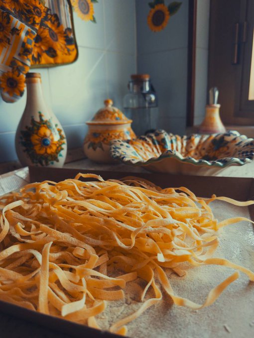 The image features a large bowl filled with pasta, placed on top of a table. The pasta appears to be made from wheat and has a yellow color that stands out against the white background. In the background, there are two vases - one containing sunflowers and another featuring a floral design. A bottle is also present in the scene, adding an element of everyday life to the image.