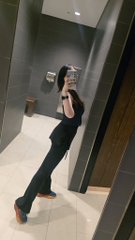 A person taking a mirror selfie in what appears to be a bathroom, wearing dark clothing.