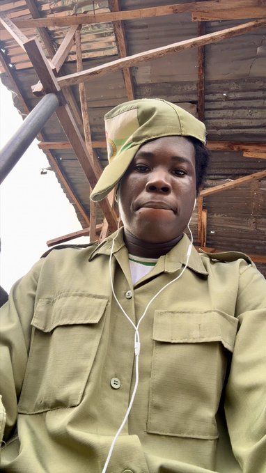 A young man in a green camouflage cap and matching uniform shirt takes a selfie while wearing white earphones, standing beneath a wooden scaffolding structure. The casual, everyday "hello" greeting pairs with his direct, confident expression as he greets the camera.