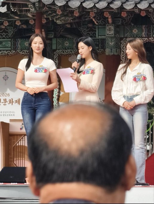 Three female hosts wearing white tops with name tags stand on stage at a 2026 Korean festival, speaking into a microphone, while the shiny bald head of a spectator in the foreground perfectly matches the post's humorous comparison to a "dead bald donkey" with an overly shiny head.