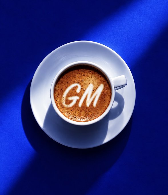 Overhead view of a white coffee cup on a saucer against a blue background, with "GM" written in foam on the coffee surface.