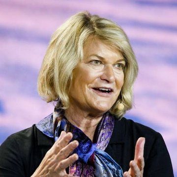 The image features a woman with blonde hair wearing a black jacket, smiling at the camera while making hand gestures. The background has a purple hue, which complements her outfit. The caption below the photo reads "Senator Cynthia Lummis: Our debt is real. Our fiscal trajectory is unsustainable. Bitcoin is one of the few tools that could help right that wrong for younger Americans." This statement seems to be related to the image's context and the woman's role as a politician, suggesting her involvement in addressing financial issues or promoting digital currency solutions.