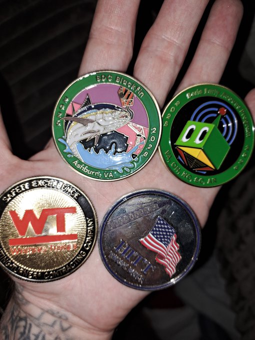 A hand displays four challenge coins: a green 'EPC Blu/AN' coin with a swordfish, a green 'Kyle Fair Autism' coin featuring a blocky green character, a gold 'WI Security' coin, and a silver coin with an American flag reading 'SEMPER FI'.