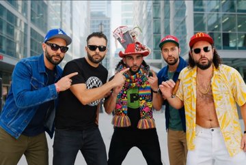 Five men pose together outdoors, with the central figure wearing an elaborate hat and colorful attire, possibly a music group photo.