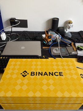 The image features a yellow box with "binance" written on it, placed on top of a desk. The desk also has various items such as a laptop computer, keyboard, mouse, cell phone, and other electronics. There are two people in the background who appear to be working or engaged in some activity related to the desk's contents.