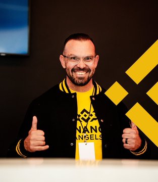 A smiling man with glasses wearing a yellow and black 'Binance Angels' jacket gives two thumbs up in front of a Binance logo wall.