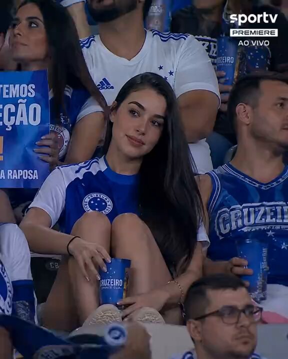 The image shows a woman sitting down, wearing blue clothing with white stars on her shirt. She has long hair and is holding a cup of beer while watching a sports game. The surrounding crowd appears to be engaged in the same activity as she is, creating an atmosphere of enjoyment and camaraderie among the fans.