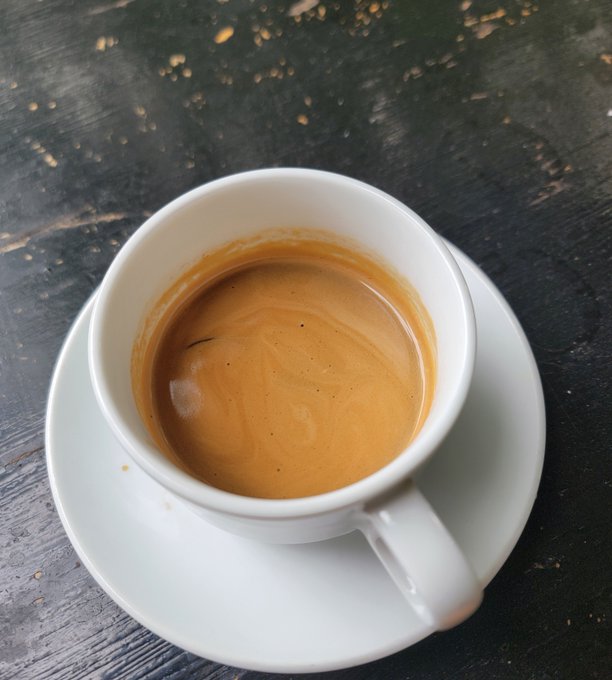 A small white espresso cup sits on a matching saucer atop a dark wooden table, filled with a freshly brewed coffee topped with a rich golden crema. The image accompanies a wish for a relaxing weekend, capturing a quiet, peaceful coffee moment to complement the sentiment of enjoying a pleasant day.