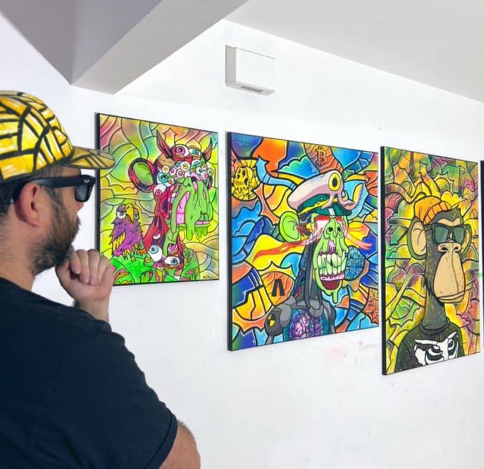 A man wearing a yellow and black checkered cap and sunglasses viewing three colorful, psychedelic-style paintings of monkeys and other creatures on a white gallery wall.