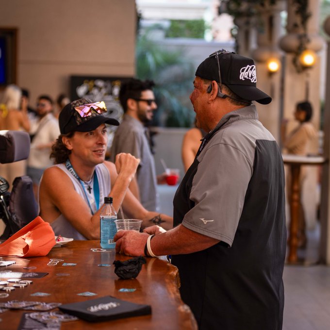 Two men at a bar or poker table engaged in conversation, one wearing a black cap and sunglasses, the other in a gray and black polo shirt, with drinks and cards visible on the counter.