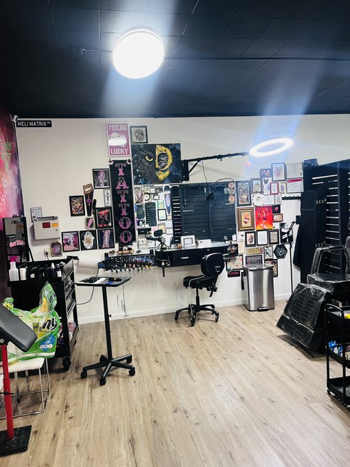 An empty tattoo/piercing studio with walls covered in framed artwork, a black desk, and a 'TATTOO' sign hanging in the center.