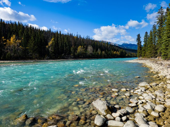 A pristine mountain river with crystal-clear turquoise waters flows through a rocky bed, bordered by dense evergreen forests and distant peaks under a vivid blue sky—capturing the essence of a fresh, vibrant "Green Morning" in nature.
