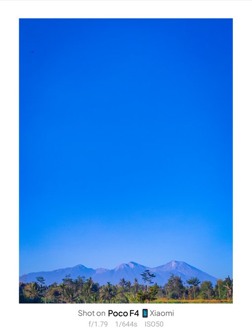 A landscape photograph showing a clear blue sky over a distant mountain range framed by green trees, shot on a Poco F4 with f/1.79, 1/644s, ISO50 settings displayed at the bottom.